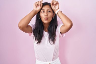 Young hispanic woman standing over pink background doing funny gesture with finger over head as bull horns 