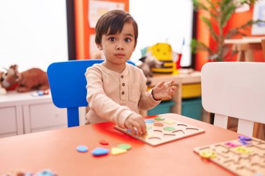 Adorable hispanic boy playing with maths puzzle game sitting on table at kindergarten