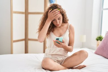 Genç, güzel, İspanyol bir kadın yatak odasında endişeli bir ifadeyle akıllı telefon kullanıyor.