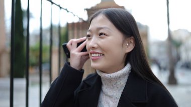 Young chinese woman smiling confident listening audio message by the smartphone at street