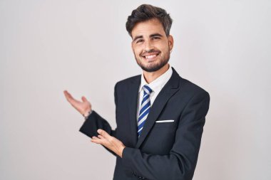 Young hispanic man with tattoos wearing business suit and tie inviting to enter smiling natural with open hand 