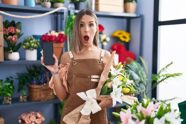 Young woman working at florist shop showing smartphone screen afraid and shocked with surprise and amazed expression, fear and excited face.