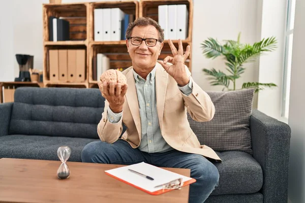 Senior psychiatrist man working at consultation office doing ok sign with fingers, smiling friendly gesturing excellent symbol