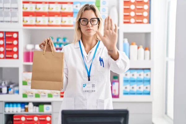 Young blonde woman working at pharmacy drugstore holding paper bag with open hand doing stop sign with serious and confident expression, defense gesture
