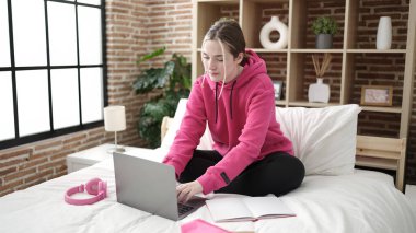 Young blonde woman student using laptop studying on bed at bedroom