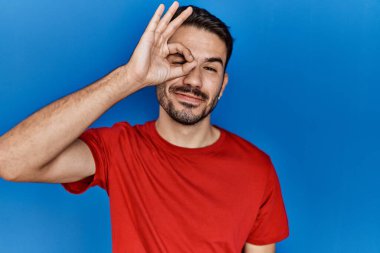 Young hispanic man with beard wearing red t shirt over blue background doing ok gesture with hand smiling, eye looking through fingers with happy face. 