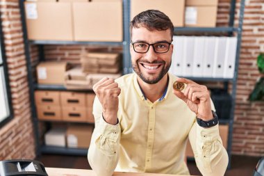 Handsome hispanic man working at small business ecommerce holding bitcoin screaming proud, celebrating victory and success very excited with raised arms 