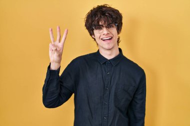 Young man wearing glasses over yellow background showing and pointing up with fingers number three while smiling confident and happy. 