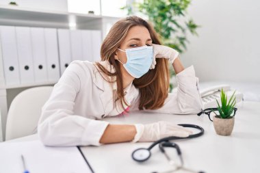 Young woman wearing doctor uniform and medical mask tired sitting on table at clinic