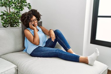 Genç Afrikalı Amerikalı kadın gülümsüyor. Kendine güveni tam. Evde akıllı telefondan konuşuyor.