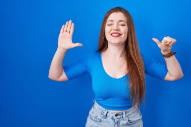 Redhead woman standing over blue background showing and pointing up with fingers number six while smiling confident and happy. 
