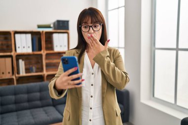 Young brunette woman working at the office with smartphone covering mouth with hand, shocked and afraid for mistake. surprised expression 