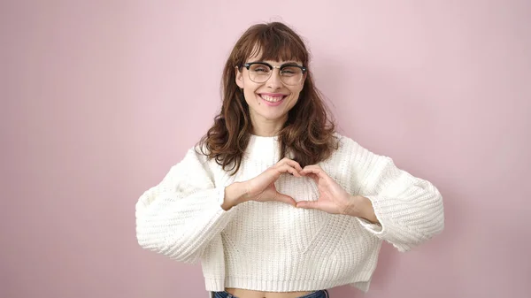 Young caucasian woman doing heart shape with hands over isolated pink background
