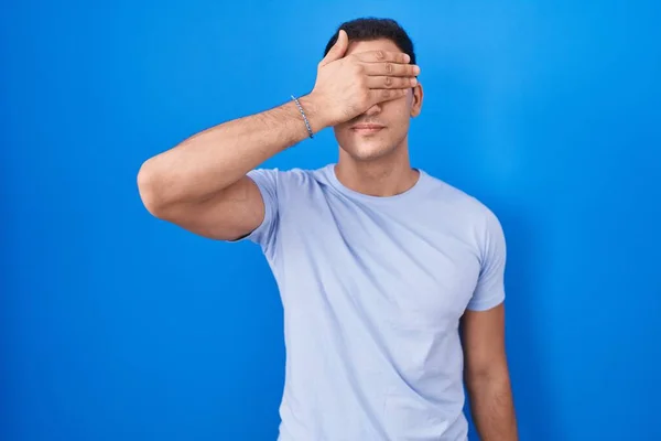 Young Hispanic Man Standing Blue Background Covering Eyes Hand Looking — Φωτογραφία Αρχείου