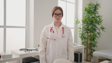Klinikte kollarını kavuşturmuş doktor üniforması giyen genç sarışın kadın.