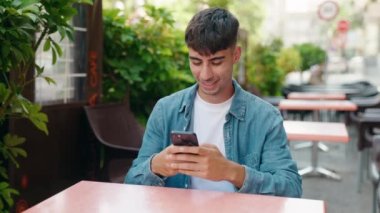 Young hispanic man using smartphone sitting on table at coffee shop terrace