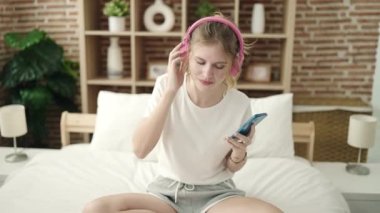 Young blonde woman sitting on bed listening to music and singing at bedroom