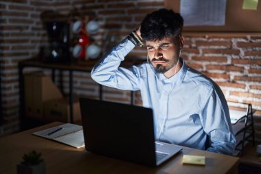 Young hispanic man with beard working at the office at night confuse and wonder about question. uncertain with doubt, thinking with hand on head. pensive concept. 