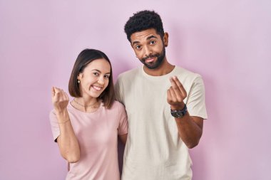 Young hispanic couple together over pink background doing money gesture with hands, asking for salary payment, millionaire business 