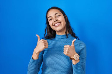 Young brazilian woman standing over blue isolated background success sign doing positive gesture with hand, thumbs up smiling and happy. cheerful expression and winner gesture. 