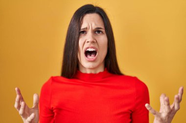 Young hispanic woman standing over yellow background crazy and mad shouting and yelling with aggressive expression and arms raised. frustration concept. 