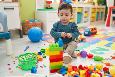 Adorable hispanic boy playing with construction blocks sitting on floor at kindergarten