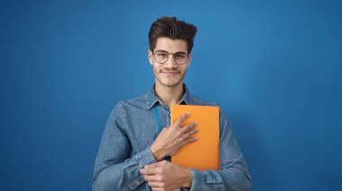 Young hispanic man smiling confident holding book over isolated blue background
