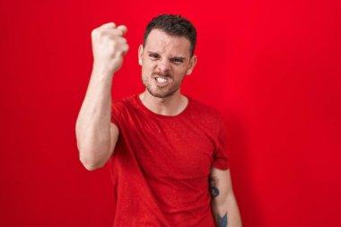 Young hispanic man standing over red background angry and mad raising fist frustrated and furious while shouting with anger. rage and aggressive concept. 