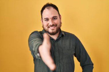 Plus size hispanic man with beard standing over yellow background smiling friendly offering handshake as greeting and welcoming. successful business. 