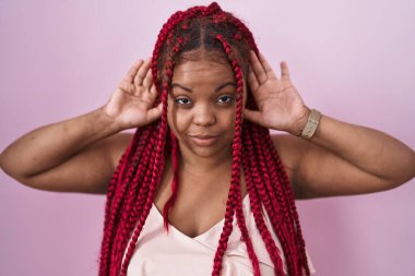 African american woman with braided hair standing over pink background trying to hear both hands on ear gesture, curious for gossip. hearing problem, deaf 