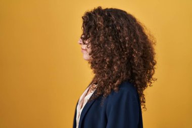 Hispanic woman with curly hair standing over yellow background looking to side, relax profile pose with natural face with confident smile. 