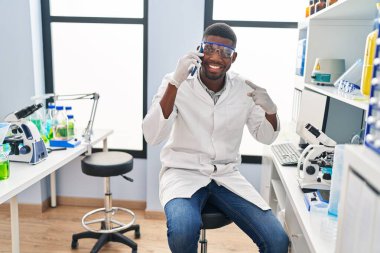 African american man working at scientist laboratory speaking on the phone smiling happy pointing with hand and finger 