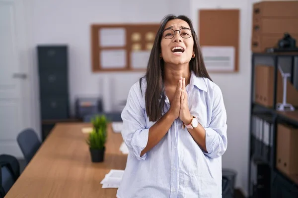 Young hispanic woman at the office begging and praying with hands together with hope expression on face very emotional and worried. begging.