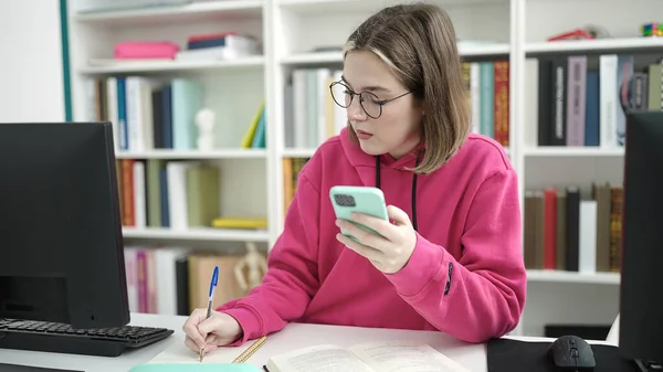 在图书馆大学 年轻的金发女学生在笔记本上使用智能手机书写 — 图库照片