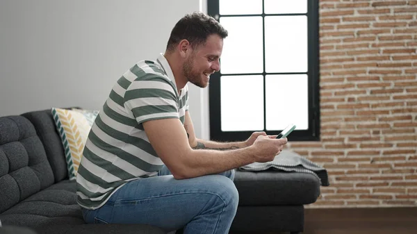 Young Caucasian Man Smiling Using Smartphone Sitting Sofa Home — Zdjęcie stockowe