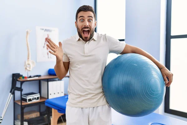 Handsome Hispanic Man Holding Pilates Ball Rehabilitation Clinic Celebrating Victory — ストック写真
