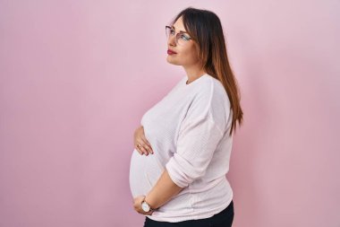 Pregnant woman standing over pink background looking to side, relax profile pose with natural face and confident smile. 