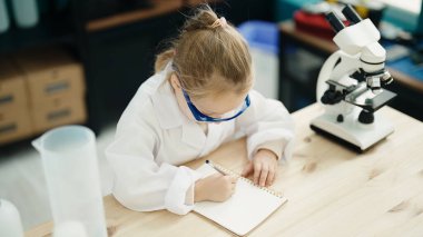 Adorable blonde girl student using microscope writing on notebook at laboratory classroom