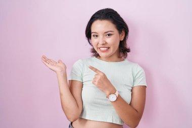 Hispanic young woman standing over pink background amazed and smiling to the camera while presenting with hand and pointing with finger. 
