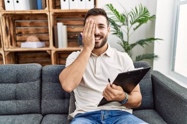 Handsome hispanic man holding clipboard working at psychology clinic covering one eye with hand, confident smile on face and surprise emotion. 