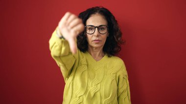 Middle age hispanic woman doing negative gesture with thumb down over isolated red background