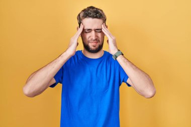 Hispanic man with beard standing over yellow background with hand on head, headache because stress. suffering migraine. 