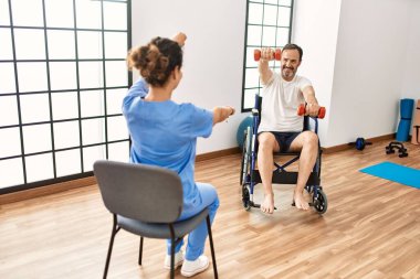 Orta yaşlı bir adam ve kadın fizyoterapi kliniğinde tekerlekli sandalyede oturan dambıllarla rehabilitasyon seansı yapıyorlar.