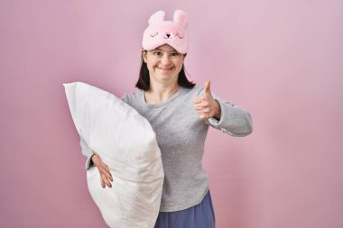 Woman with down syndrome wearing sleeping mask hugging pillow doing happy thumbs up gesture with hand. approving expression looking at the camera showing success. 