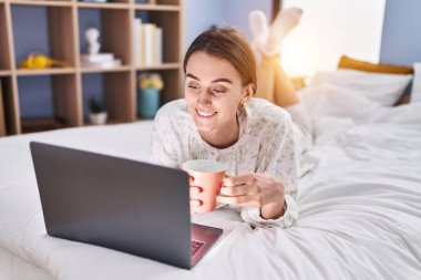 Young caucasian woman using laptop drinking coffee at bedroom