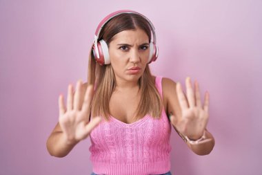 Young blonde woman listening to music using headphones moving away hands palms showing refusal and denial with afraid and disgusting expression. stop and forbidden. 