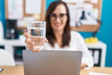 Young beautiful hispanic woman business worker using laptop drinking water at office
