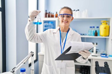 Young blonde woman wearing scientist uniform holding test tube and clipboard at laboratory