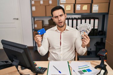 Young hispanic man with beard working at small business ecommerce holding banknotes and credit card in shock face, looking skeptical and sarcastic, surprised with open mouth 