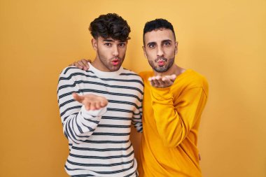 Young hispanic gay couple standing over yellow background looking at the camera blowing a kiss with hand on air being lovely and sexy. love expression. 